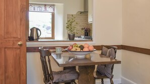 A dining area with a wooden table and chairs, a bowl of fruit, and a window with a view of houses. - 1 bedroom accommodation in Lostwithiel (Lostwithiel)