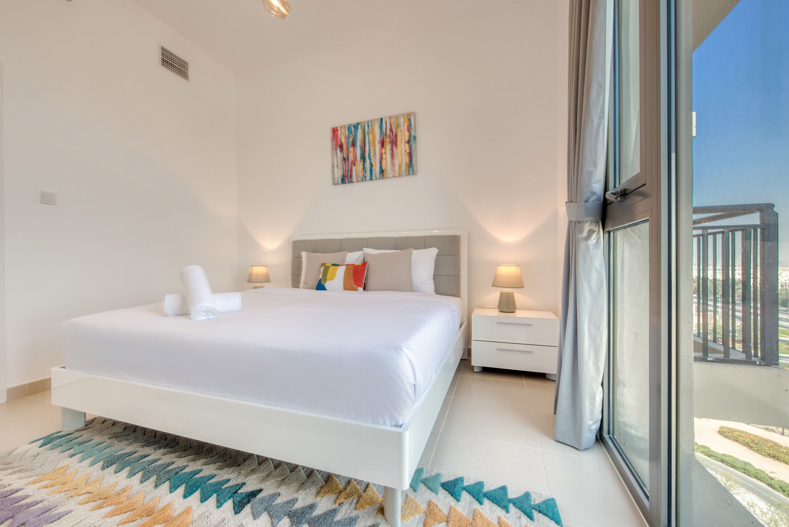 A modern bedroom with a large bed, bedside tables with lamps, a colorful abstract painting, and a balcony with a railing.