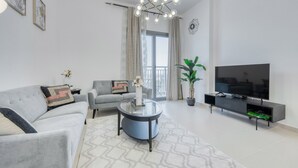 En moderne stue med en grå sofa, et rundt sofabord, et fladskærms-tv på et sort stativ og et stort vindue med gardiner.