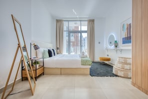 Ein modernes Schlafzimmer mit einem großen Bett, einem Nachttisch mit Lampe, einem kleinen Hocker, einem Spiegel und einer dekorativen Vase.