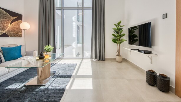 Ett modernt vardagsrum med ett soffbord i glas, en soffa med blå kuddar, en krukväxt, en platt-TV som är monterad på väggen och ett stort fönster med grå gardiner.