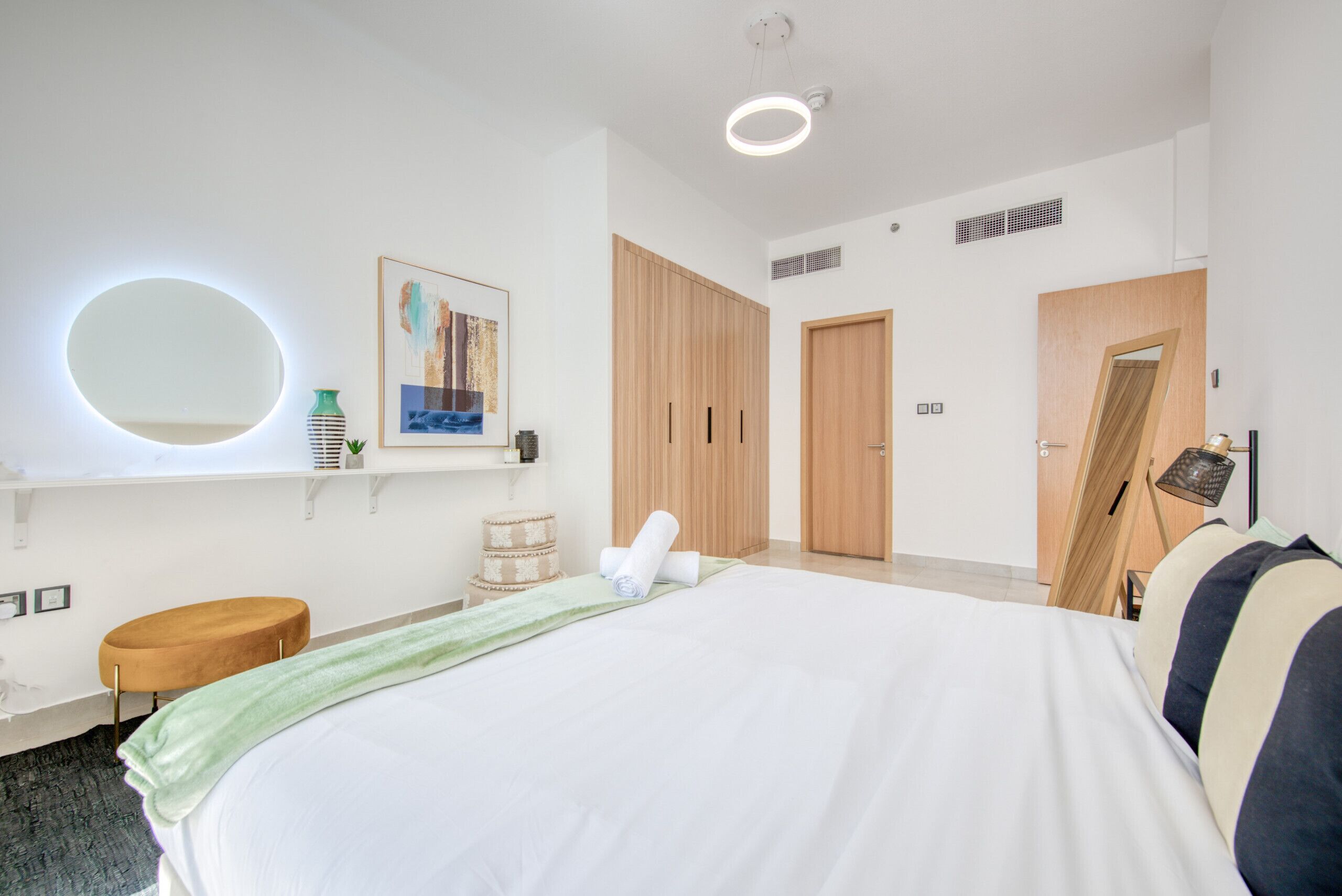Una habitación de hotel moderna con una cama grande, un espejo redondo, un armario de madera y una puerta que da a otra habitación.