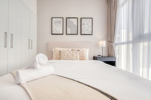 A neatly made bed with a beige blanket, a rolled towel, and a pillow with a floral pattern. There are three framed artworks on the wall above the bed.
