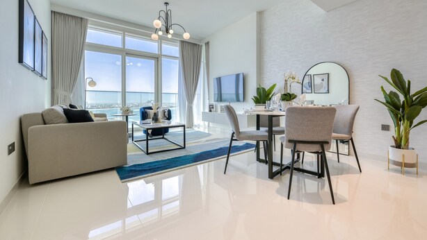 A modern living room with a sofa, dining table, chairs, and a view of the city. - Idyllic 1BR at Sunrise Bay Tower 1 Emaar Beachfront Dubai Marina by Deluxe Holiday Homes (Dubai)