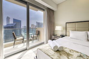 A hotel room with a large balcony, a bed with white linens, a nightstand with a lamp, and a view of the city skyline.