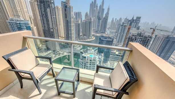 A balcony with two wicker chairs and a small table, overlooking a cityscape with high-rise buildings and a marina. - Pristine 3BR at The Address Residences Dubai Marina by Deluxe Holiday Homes (Dubai)