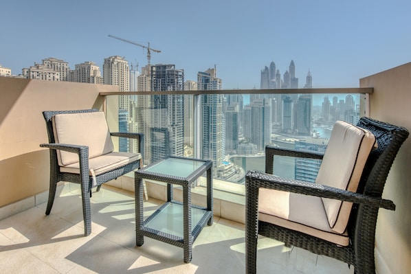 A balcony with wicker furniture and a glass table, overlooking a cityscape with high-rise buildings and a construction crane.