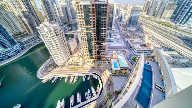 A high-rise building with a modern design, surrounded by other tall buildings, a marina with boats, and a swimming pool.