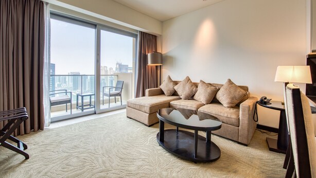 A modern hotel room with a large window, a sofa, a coffee table, and a desk with a lamp.