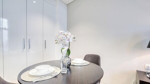 Un coin repas avec une table ronde dressée pour deux personnes, un vase rempli de fleurs blanches et, au fond, une cuisine moderne.
