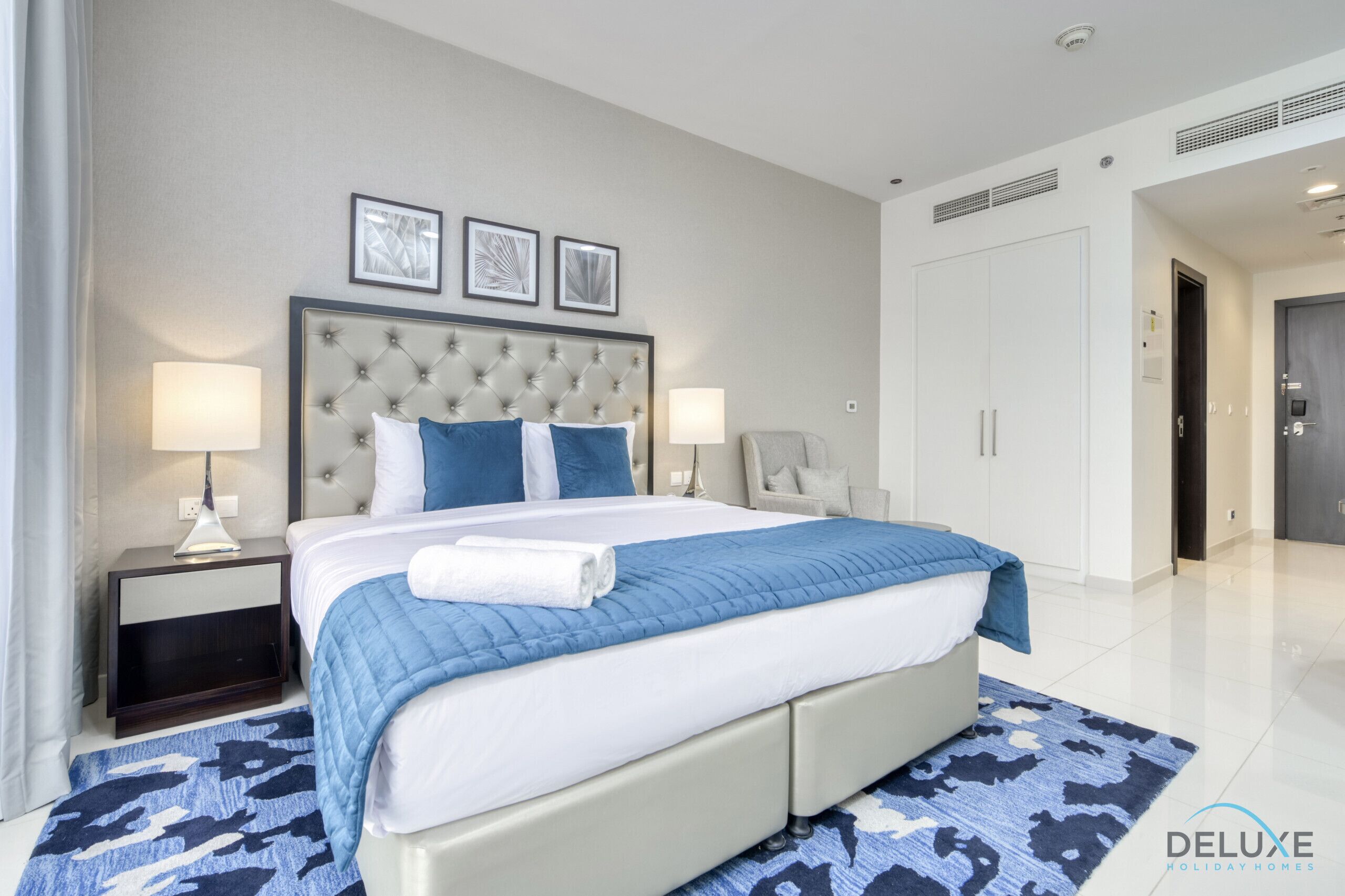 A modern hotel bedroom with a large bed, two bedside tables with lamps, a wardrobe, and a door leading to another room.