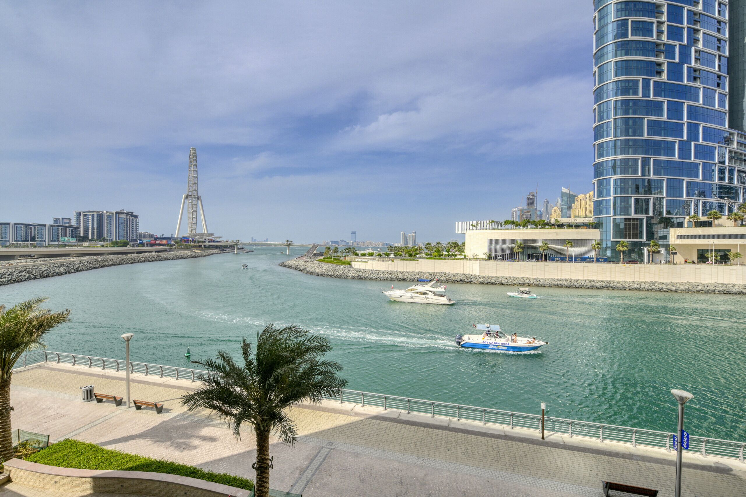 Eine Uferpromenade mit einem modernen Gebäude, einem Riesenrad und Booten auf dem Wasser.