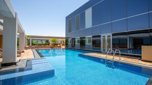 A modern hotel with a swimming pool, sun loungers, and a covered area with a palm tree.