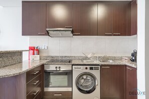 A modern kitchen with dark wood cabinets, a stainless steel range hood, a built-in oven, and a front-loading washing machine. - Sunny Studio at Sky Gardens DIFC by Deluxe Holiday Homes (Dubai)