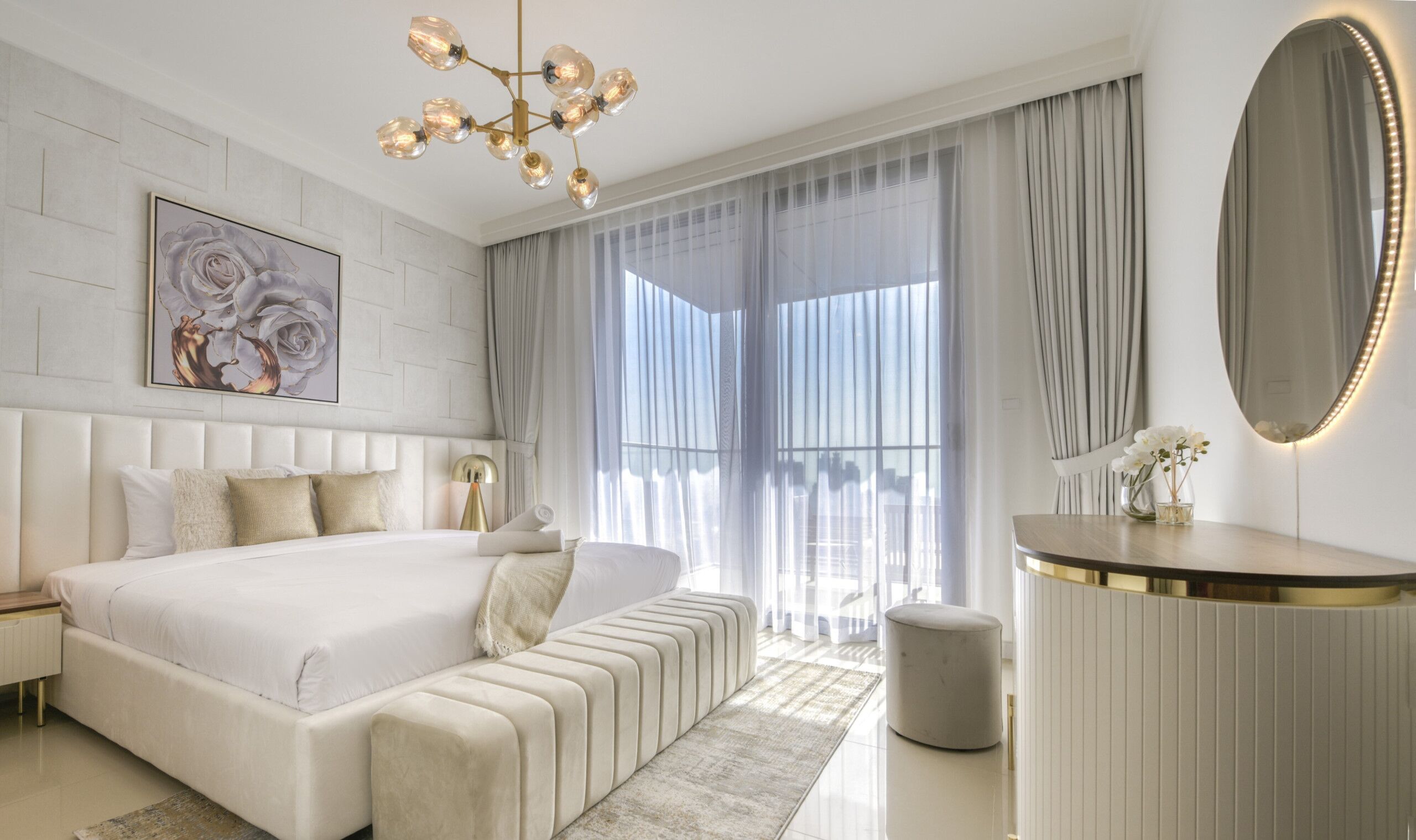 Une chambre moderne avec un grand lit, une table de chevet, une coiffeuse ronde avec un miroir et une grande fenêtre ornée de rideaux légers.