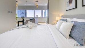 Moderni hotellihuone, jossa on suuri sänky, yöpöytä, tuoli ja suuret ikkunat, joista avautuu näkymä kaupunkiin.