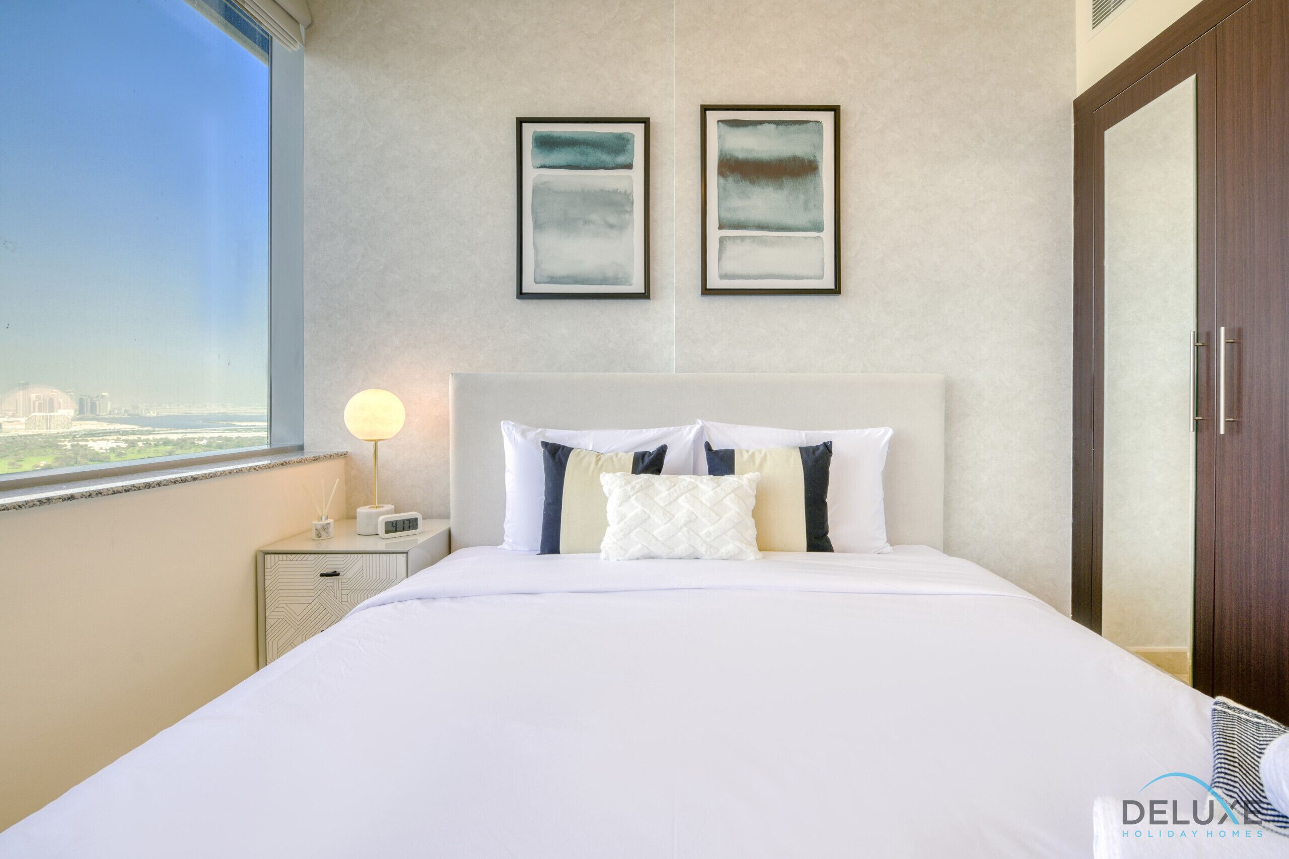 A hotel room with a large bed, a nightstand with a lamp, two framed artworks on the wall, and a view of the city through the window.