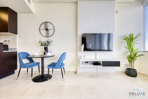Una sala de estar moderna con una mesa redonda, dos sillas azules, un televisor montado en la pared, una planta en maceta y una zona de cocina con armarios oscuros.