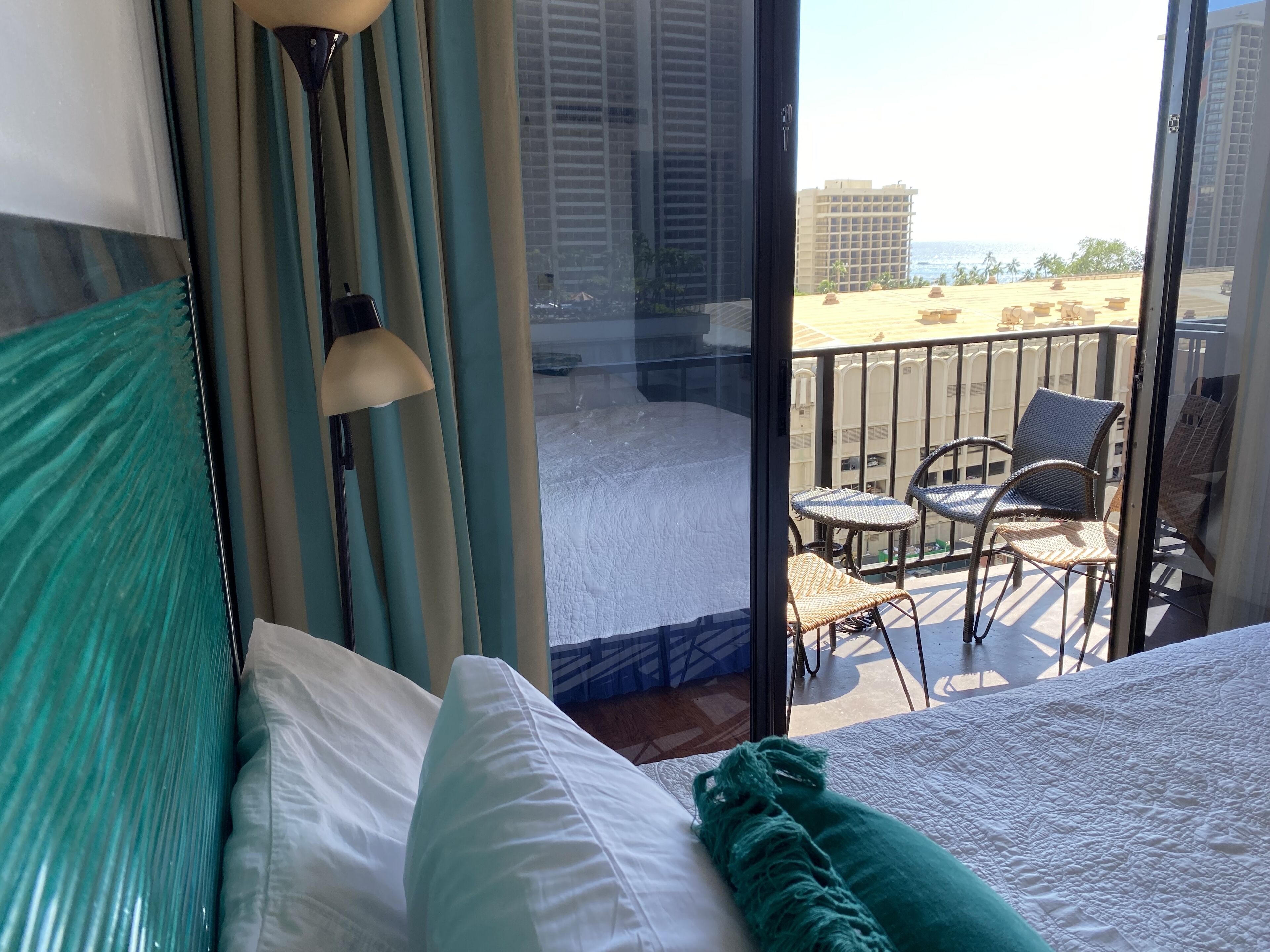 A hotel room with a bed, a window with curtains, a balcony with chairs, and a view of buildings and the sea.