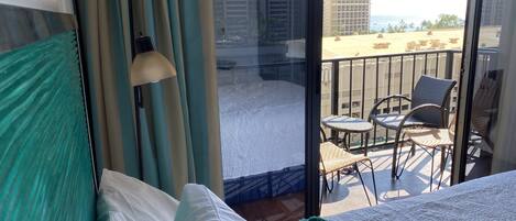 A hotel room with a bed, a window with curtains, a balcony with chairs, and a view of buildings and the sea.