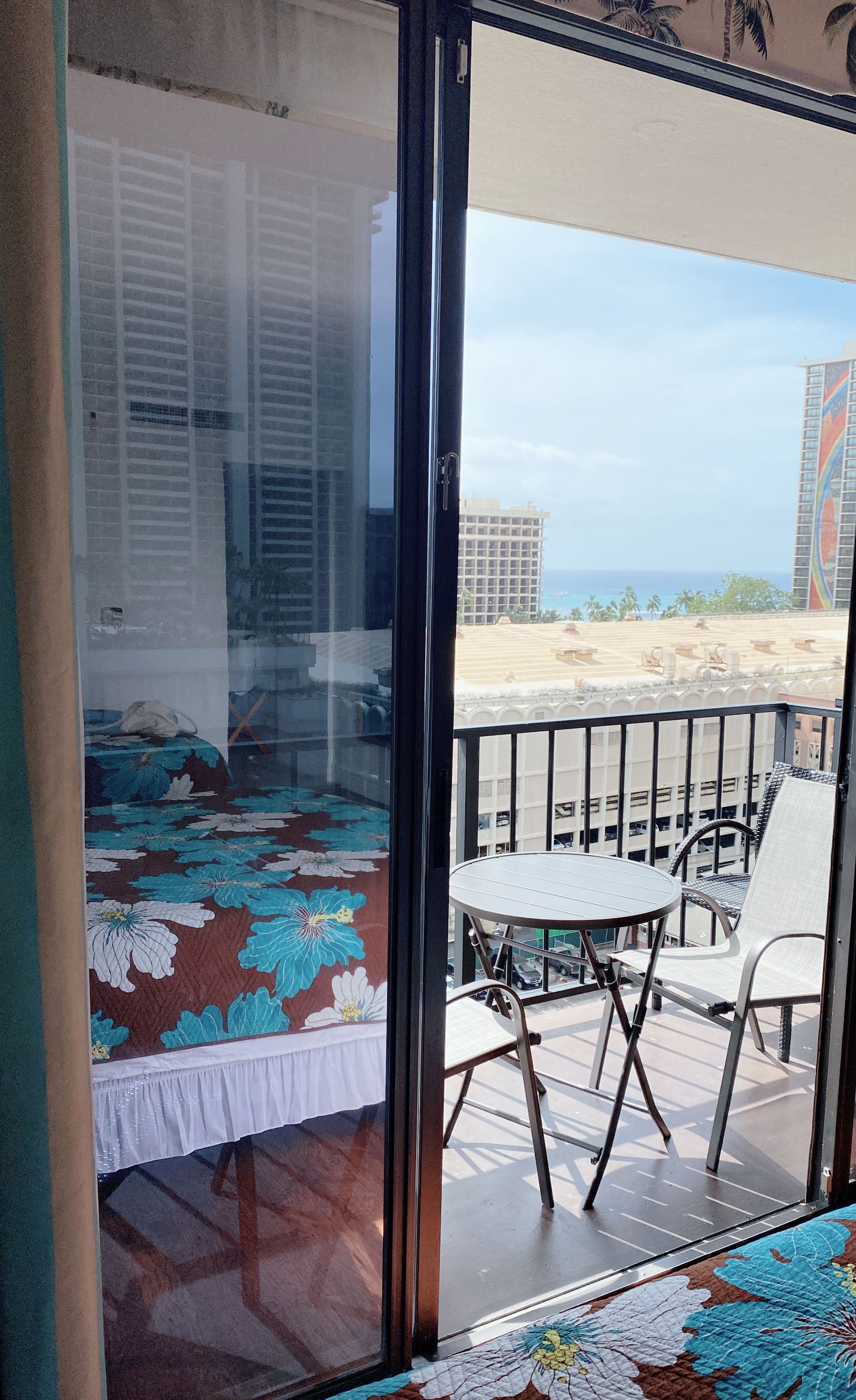 A balcony with a view of the beach and buildings, a bed with a floral bedspread, and a table with chairs.