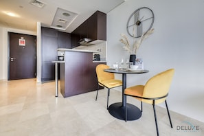 A modern hotel room with a dining area featuring a round table and two yellow chairs, a kitchen area, and a decorative wall clock.
