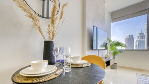 Un área de comedor con una mesa redonda dispuesta para dos personas, un jarrón con plantas secas y vistas a un paisaje urbano a través de un ventanal grande.