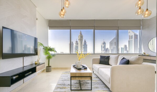 Uma sala de estar moderna com uma televisão grande, um sofá, uma mesa de centro e vista para o horizonte urbano através de grandes janelas.