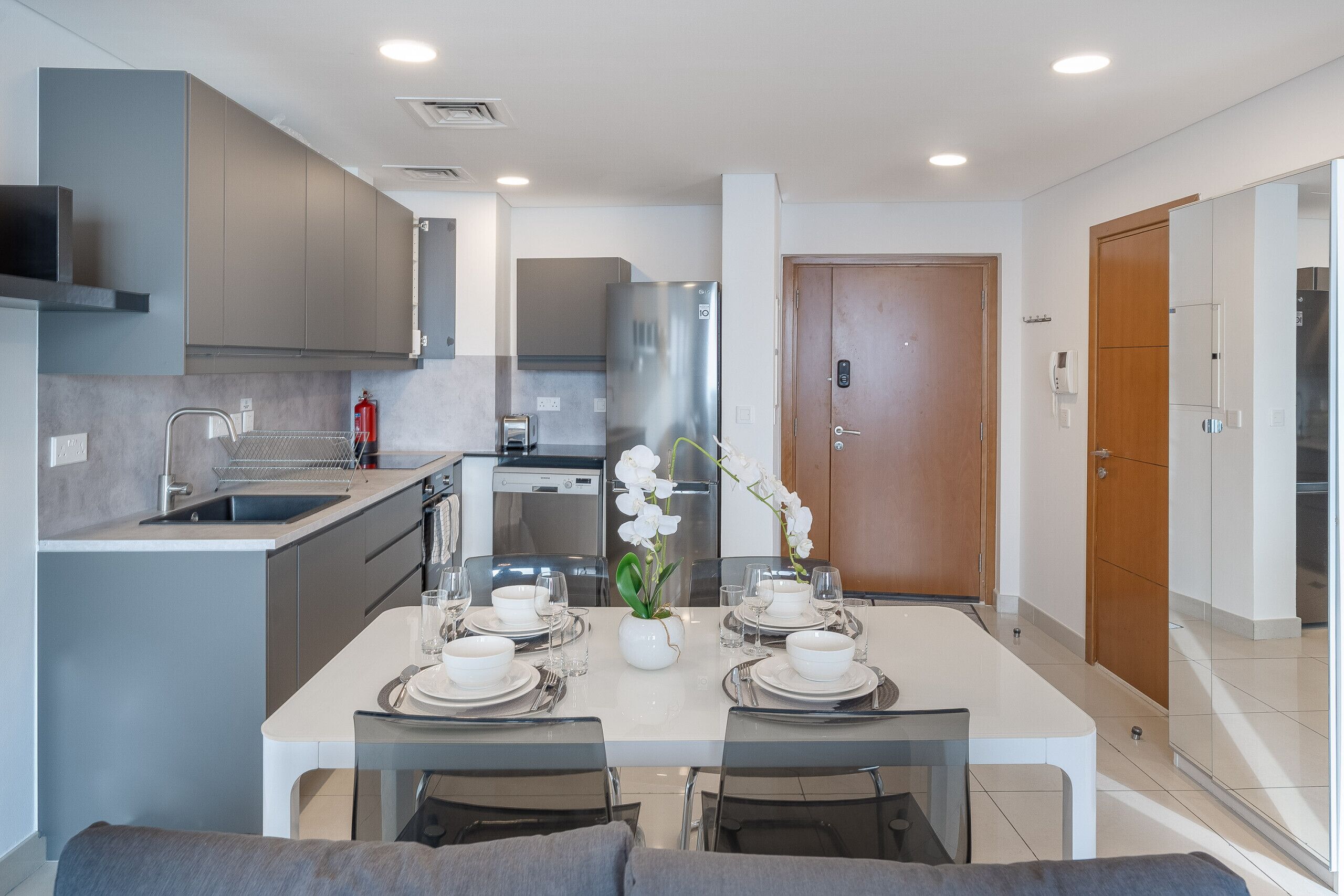A modern kitchen with grey cabinets, a stainless steel refrigerator, and a dining area set for four with white tableware.