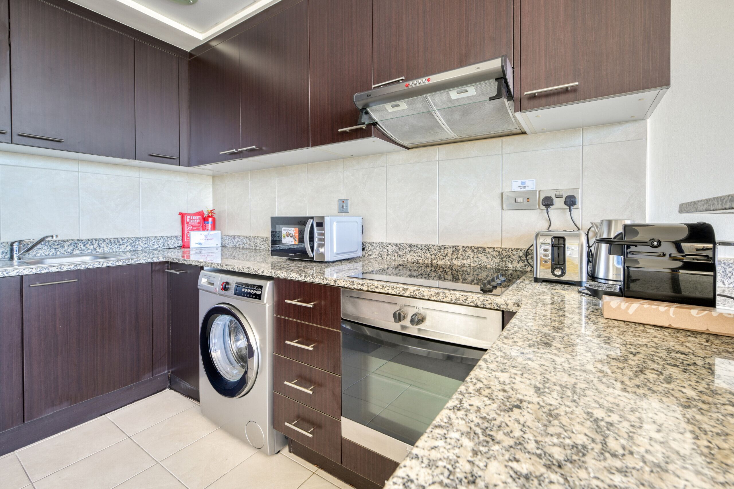 A modern kitchen with dark wooden cabinets, a stainless steel range hood, a built-in washing machine, a microwave, a toaster, and a coffee maker.