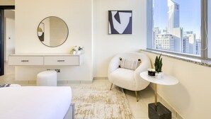 Quarto de hotel moderno com uma janela grande, um espelho redondo, uma poltrona branca, uma pequena mesa redonda com uma planta e um design minimalista.