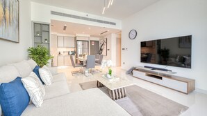Una sala de estar moderna con sofá, mesa de centro y televisor. Se observa una cocina al fondo con armarios y comedor.