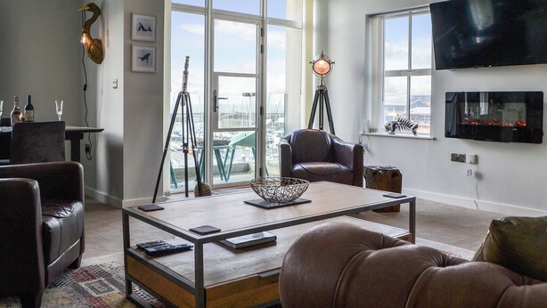 Een moderne woonkamer met een groot raam, een hangende televisie en een eethoek met een tafel en stoelen.