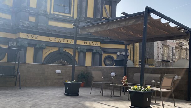 Un bâtiment portant l’inscription « AVE GRATIA PLENA », avec des tables et des chaises à l’extérieur, ainsi que des plantes en pot.