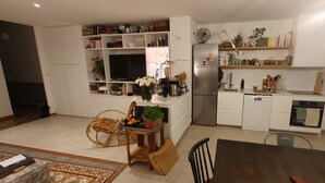 Una cocina moderna con armarios blancos, horno integrado y refrigerador. Cuenta con una mesa de comedor con sillas y una silla de mimbre. La cocina tiene estantes con diversos objetos y una planta en maceta.