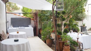 Un patio al aire libre con mesas redondas cubiertas con manteles blancos, rodeado de plantas en macetas y una valla de madera.