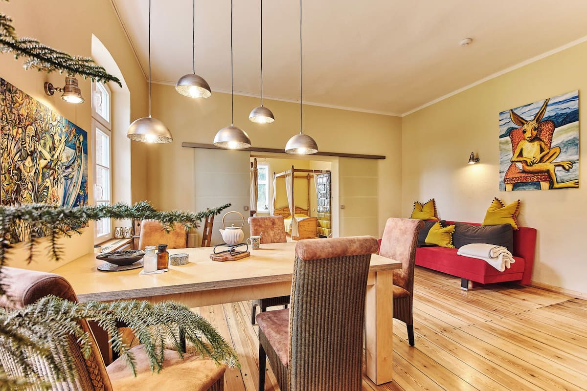 Een gezellige woonkamer met een houten vloer, een rode bank met gele kussens, een eethoek met een houten tafel, en een groot schilderij aan de muur.