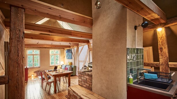A rustic interior with exposed wooden beams, a brick fireplace, and a dining area with wooden flooring and a table. - Kulturhof Dretzen (Buckautal)