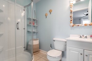 A bathroom with a glass shower enclosure, a white toilet, a sink with a mirror, and shelves with toiletries.