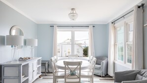 A dining area with a round table, white chairs, a white cabinet with a mirror, and a view of houses through a large window. - Carolina House - Steps to Beach, Fantastic Location!  Walk to All Attractions (Emerald Isle)