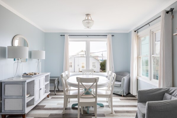 A dining area with a round table, white chairs, a white cabinet with a mirror, and a view of houses through a large window.