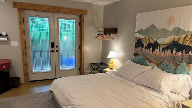 A bedroom with a bed, a nightstand, a lamp, a wall-mounted coat rack, and a door with frosted glass panels. - Private apartment in a quiet neighborhood with lots of trees and privacy. (Weaverville)