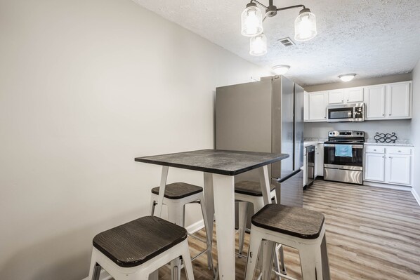 A compact kitchen with white cabinets, a microwave, a stove, and a dining area with a table and stools. - Grand Park, Westfield, Space For Entire Team, Sleeps 30 comfortably. Unit B (Sheridan)