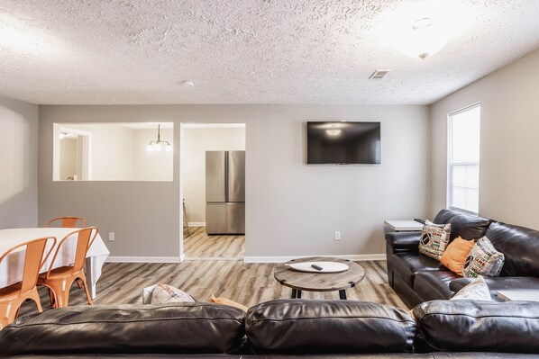 Una sala de estar moderna con un sofá de cuero negro, un comedor con sillas color naranja, una cocina con electrodomésticos de acero inoxidable y un televisor de pantalla plana montado en la pared.