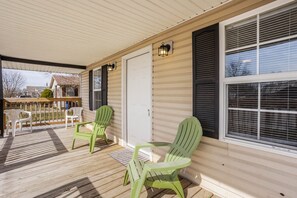 A covered porch with wooden flooring, two green Adirondack chairs, a white door, and a window with blinds. - Grand Park, Westfield, Space For Entire Team, Sleeps 30 comfortably. Unit B (Sheridan)