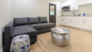 A modern living room with a grey sectional sofa, a patterned ottoman, a textured coffee table, and a kitchen area with white cabinets and a microwave. - Cozy Lake House: Unique Japanese Garden Stay in Miami (Homestead)