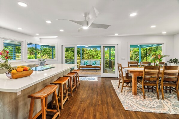 A modern kitchen with a central island, wooden bar stools, and a dining area with a large table and chairs. - Hoaloha- Few Steps to Hanalei Bay+AC & Hot Tub (Hanalei)