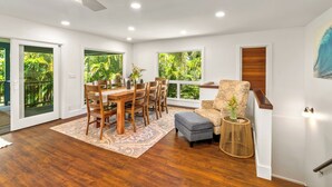 Habitación amplia con una mesa de comedor de madera, sillas y un tapete estampado. Se observa una ventana con vista a follaje, un ventilador de techo y una pequeña mesita con una planta.
