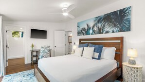 Un dormitorio con una cama grande, un ventilador de techo, un televisor montado en la pared y un cuadro de palmeras.
