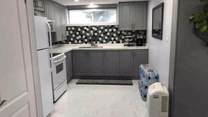 A modern kitchen with grey cabinets, a white refrigerator, a stove, and a microwave. There is a window above the sink and a plant on the right. - Beautiful One bedroom basement apartment (Toronto)